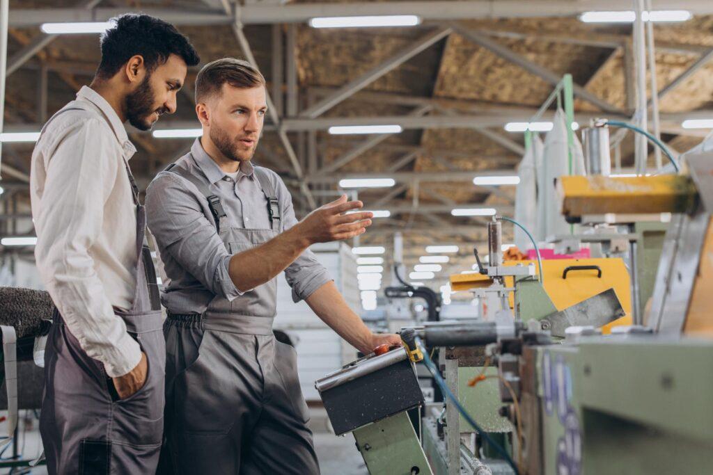Ein männlicher Inspektor oder Bediener einer Werkstatt für die Herstellung von Aluminium- und Kunststoffkränzen weist einen Praktikanten ein. Ein internationales Team von Männern arbeitet gemeinsam an einer Maschine in einer Fabrik.