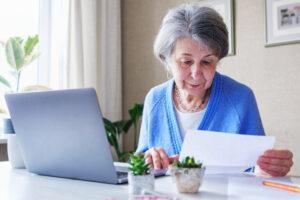Ältere Frau liest eine Mitteilung oder ein Dokument, arbeitet von zu Hause aus mit Laptop und Papierkram - Planung des Erbes oder der Altersversorgung und Verwaltung der Finanzen 