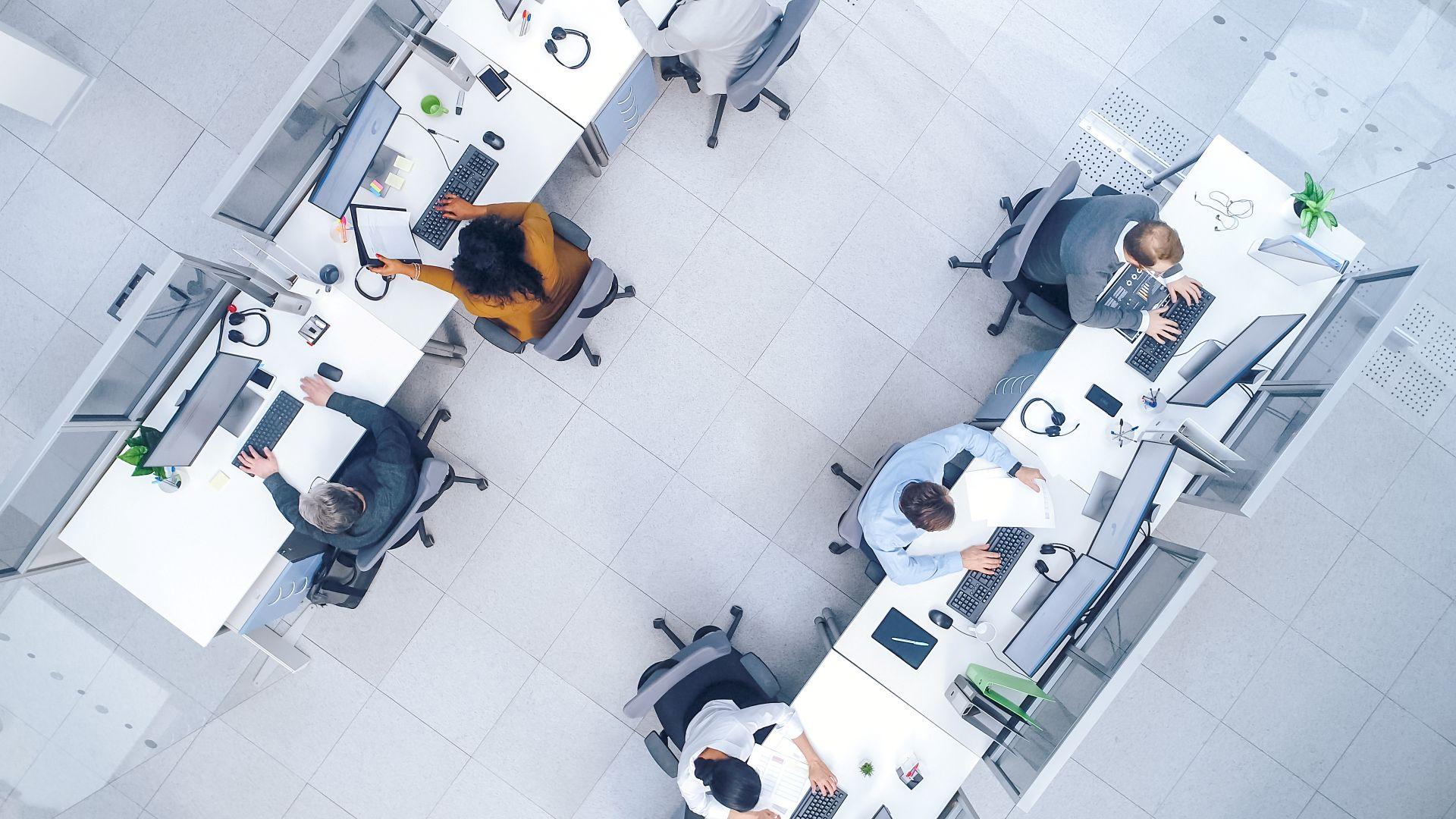 Blick von oben auf eine strukturierte Arbeitsumgebung mit mehreren Mitarbeitenden an vernetzten Schreibtischen